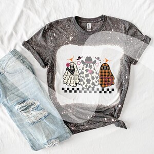 May include: A gray and white bleached t-shirt featuring a graphic of three cartoon ghosts wearing cowboy hats and bandanas. The ghosts are in different patterns, including cow print and plaid. Light blue ripped jeans are next to the shirt.