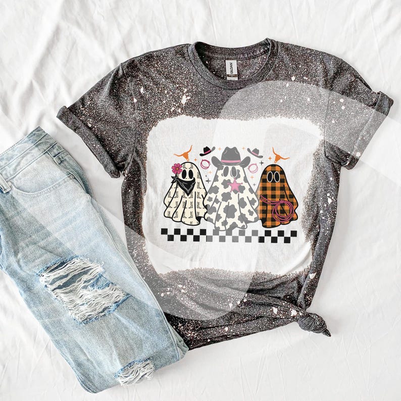 May include: A gray and white bleached t-shirt featuring a graphic of three cartoon ghosts wearing cowboy hats and bandanas. The ghosts are in different patterns, including cow print and plaid. Light blue ripped jeans are next to the shirt.
