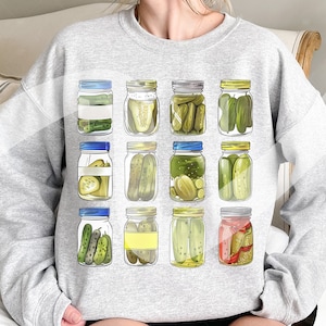 Peut inclure: Un sweat-shirt gris avec un motif de 10 bocaux de cornichons. Chaque bocal a un couvercle bleu et contient des cornichons verts.