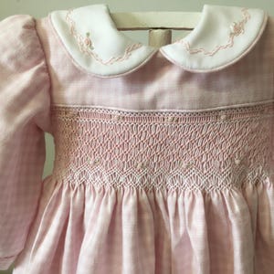 May include: A pink and white gingham dress with a white collar. The dress features a detailed smocked bodice and puffed sleeves. The collar is trimmed with pink embroidery, and the dress has a gathered skirt.