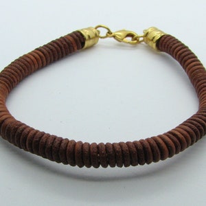 Könnte beinhalten: Ein braunes Lederarmband mit einem goldenen Verschluss. Das Armband besteht aus kleinen, runden Perlen, die miteinander verwoben sind.