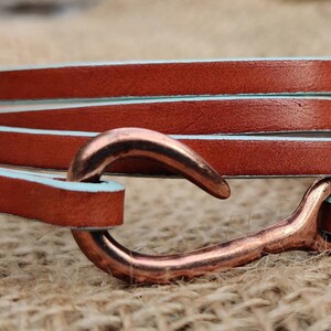 May include: A brown leather bracelet with a copper hook clasp. The leather is wrapped around three times and has a light blue edge.