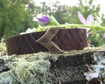May include: A brown leather bracelet with a chevron pattern and a brass arrowhead clasp. The bracelet is resting on a tree stump with moss and lichen growing on it.