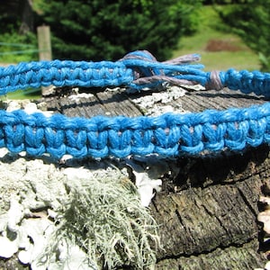 May include: A blue and gray braided bracelet made of cord. The bracelet is lying on a piece of wood with white lichen.