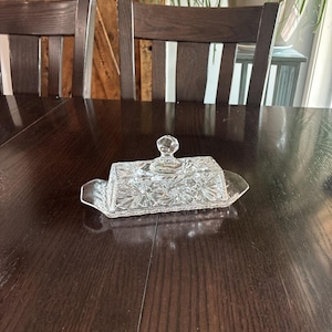 May include: A clear pressed glass butter dish featuring a rectangular base with handles and a matching lid. The glass has a faceted starburst pattern throughout and a small, round, diamond-cut knob on top of the lid for easy lifting and handling.