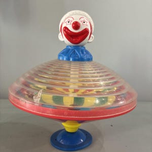 May include: A vintage plastic spinning top toy featuring a clown face on top. The top has a clear, ridged body with red, yellow, and green accents. The clown has a white face, red nose and mouth, and star-shaped eyes.