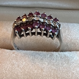 May include: A silver-toned ring featuring a multi-row design with a central band of clear, round-cut stones flanked by two rows of deep pink, round-cut stones. The stones are set in a pronged metal gallery that extends along the top of the band.
