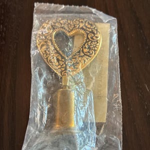 May include: A gold-toned metal lamp finial featuring a heart-shaped top with an intricate embossed floral pattern. The finial is encased in a clear plastic bag with a small paper insert visible behind it, showing the text GOLD-TONE-0.109.