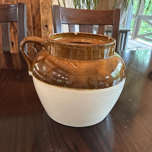 May include: A two-toned stoneware crock featuring a glossy brown glazed upper section and a matte off-white lower base. This round vessel includes a single curved handle attached to the side and a wide, open top rim with a small chip on the edge.