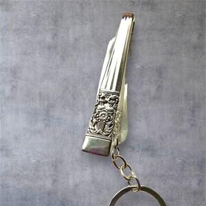 Puede incluir: Un llavero de plata con un dije de cuchara decorativa. La cuchara tiene un diseño floral y está unida a una cadena de plata con un anillo para llaves.