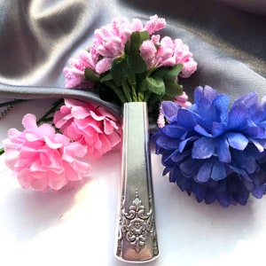May include: A silver-toned metal handle with an ornate embossed floral pattern at the base, serving as a vase for a small bouquet of artificial pink flower buds and a single large, deep blue fabric flower with layered petals.