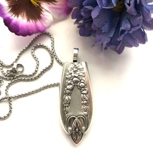 Op de afbeelding: Een zilveren ketting met een hanger gemaakt van een vintage lepel. De lepel is versierd met een bloemenmotief en heeft een hartvormige opening aan de bovenkant.