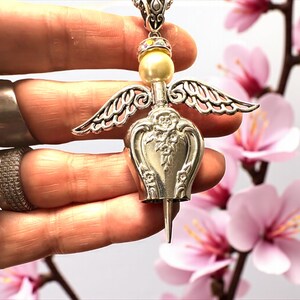 May include: A silver-toned angel pendant necklace. The pendant features a pearl bead head, ornate wings, and a decorative body. The necklace is held by a hand with rings. Pink flowers are in the background.