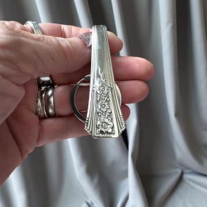 Könnte beinhalten: Silberfarbener Schlüsselanhänger mit floralem Design. Der Schlüsselanhänger wurde aus einem recycelten Löffel gefertigt. Der Griff des Löffels weist ein geprägtes Blumenmuster auf. Der Schlüsselring ist am Löffelgriff befestigt.