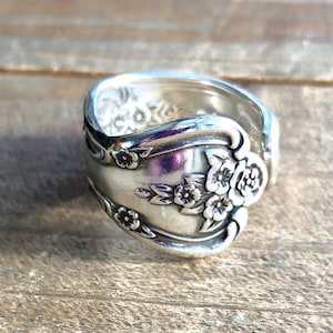 Peut inclure: Une bague en argent fabriquée à partir d'une cuillère, ornée de motifs floraux. La bague présente un design incurvé avec des fleurs et des feuilles en relief, lui donnant un aspect vintage. La bande de la bague mesure environ 1,9 cm de large.