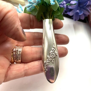 May include: A silver spoon with a floral design has been repurposed into a small vase.  A bouquet of blue and white flowers is arranged in the spoon vase.