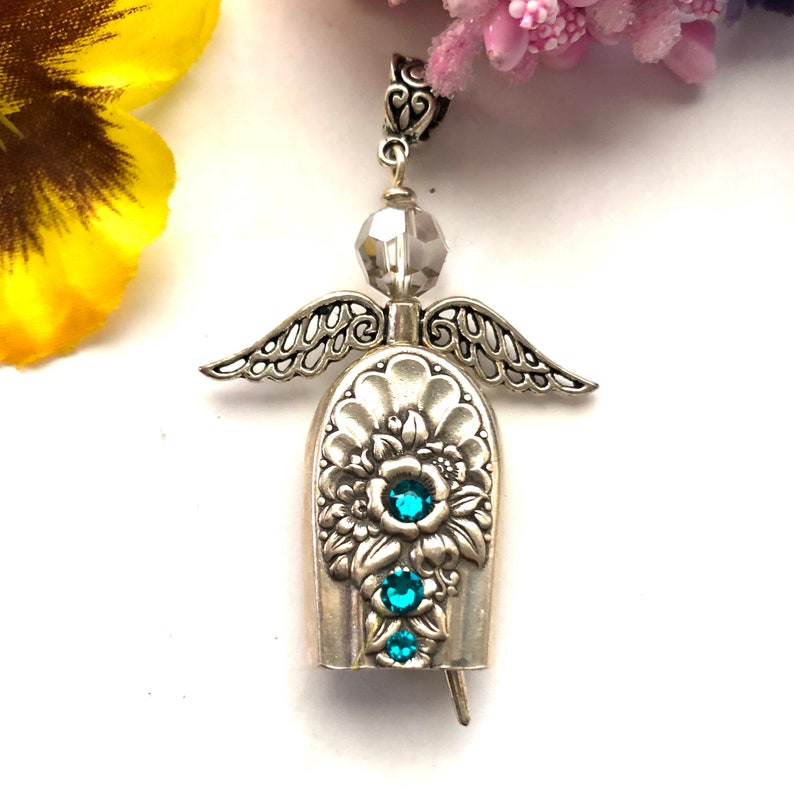 May include: A silver-toned angel pendant with intricate wing and floral details. The pendant features three teal-colored gemstones and a faceted gray bead. The angel's bell-shaped body is adorned with floral patterns.