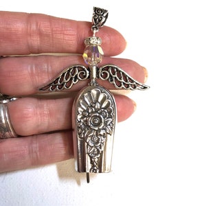 May include: A handcrafted silver angel pendant. The angel features detailed wings, a floral design on the body, and a sparkling crystal bead. The pendant is made from repurposed silverware, showcasing a unique, vintage aesthetic.