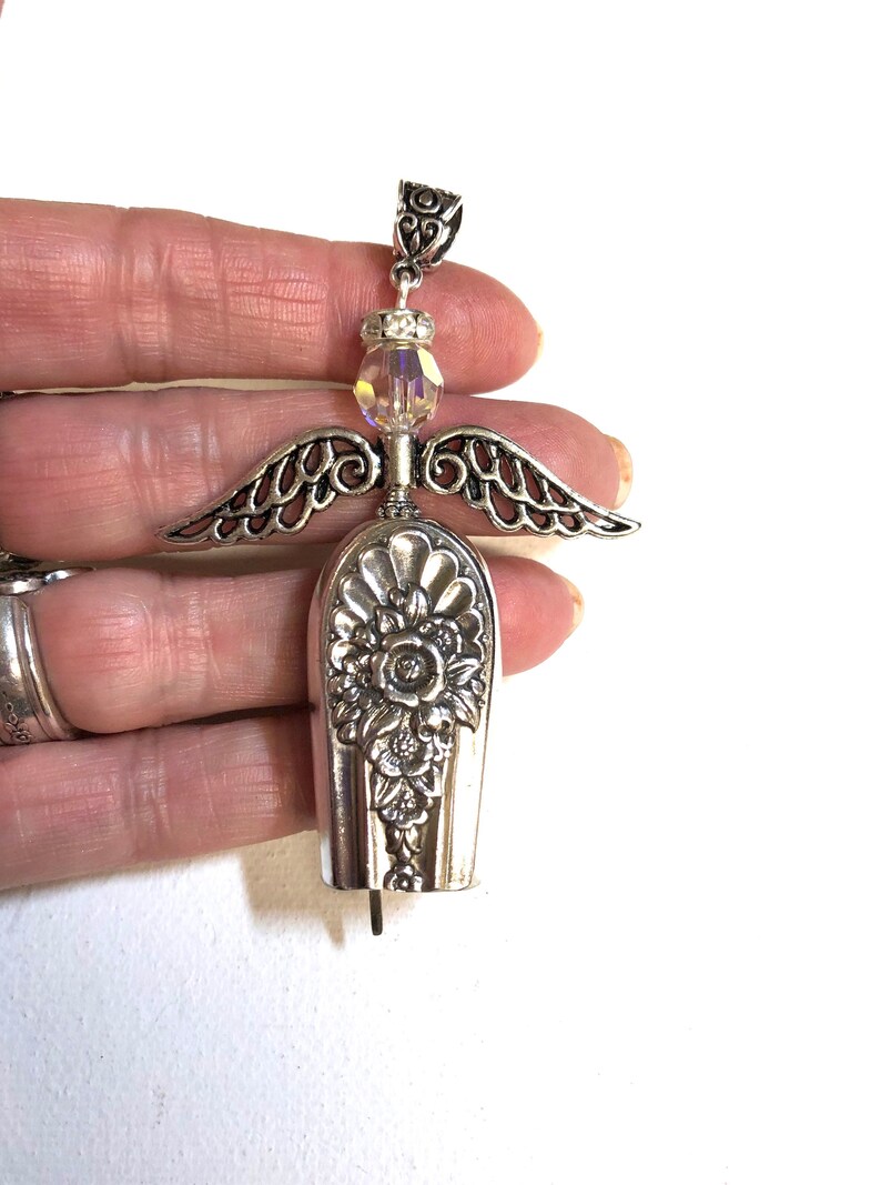 May include: A handcrafted silver angel pendant. The angel features detailed wings, a floral design on the body, and a sparkling crystal bead. The pendant is made from repurposed silverware, showcasing a unique, vintage aesthetic.