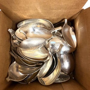May include: A collection of numerous silver-toned metal spoons with leaf-shaped bowls and stems, packed together inside a plain brown cardboard box. The spoons feature a smooth, reflective finish and are arranged in a jumbled, overlapping pile.