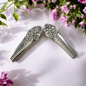 May include: Two silver hair slides with floral patterns. The slides are shaped like spoons, with detailed rose and flower designs on the top. The background is white with pink flowers and green leaves.