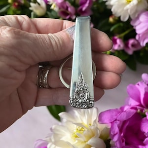 May include: A silver spoon keyring with a floral pattern at the base. The spoon is held against a blurred backdrop of pink and white flowers. The keyring loop is visible.