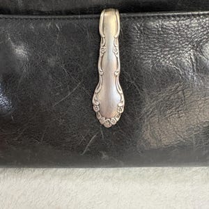 Puede incluir: Bolso de mano de cuero negro con un cierre plateado en forma de cuchara. El cierre está adornado con detalles florales y de volutas. El cuero tiene un aspecto texturizado.