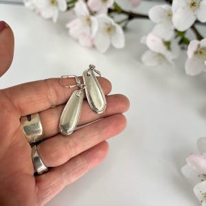 May include: Silver teardrop earrings with a lever-back clasp. The earrings have a spoon-like design with a decorative edge. The earrings are displayed against a white background with white flowers.