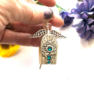 May include: A silver-toned angel pin with intricate floral detailing and three teal-colored gemstones. The angel has delicate filigree wings and a crystal bead at the top. A unique, handcrafted jewelry piece.