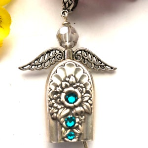 May include: A silver angel pendant with intricate wing details and a floral design on the body. The pendant features three teal-colored gemstones and a faceted gray bead at the top. The angel is suspended from a decorative silver bail.