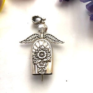May include: A silver-toned angel pendant with a floral design on the body. The pendant features delicate wings, a crystal bead head, and a loop for hanging. The angel's body is bell-shaped, with a detailed flower pattern.