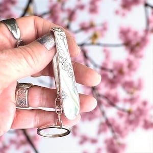 May include: A silver spoon keychain with floral and leaf patterns. The spoon is bent and attached to a keyring. The background is a soft focus of pink flowers.