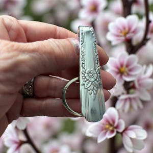 Könnte beinhalten: Ein silberfarbener Schlüsselanhänger aus einer wiederverwerteten Gabel. Der Griff der Gabel hat ein geprägtes Blumendesign und vertikale Linien. Der Schlüsselanhängerring ist am Griff der Gabel befestigt. Der Hintergrund ist verschwommen mit rosa und weißen Blüten.