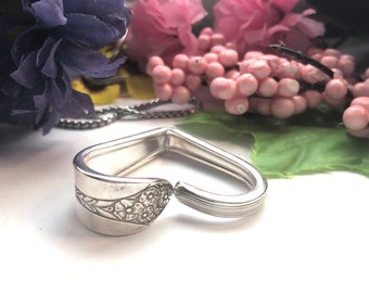 Puede incluir: Un brazalete de plata en forma de corazón hecho de una cuchara reutilizada. La cuchara tiene un diseño floral en un lado y un patrón texturizado en el otro.