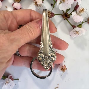 May include: Silverware keychain made from a vintage spoon. The spoon features ornate floral detailing and a key ring. The keychain is held against a backdrop of white and pink flowers.