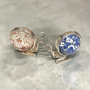 May include: Two decorative snail figurines crafted from silver-toned metal forks. One snail features a clear glass shell with red flecks, while the other has a blue and white speckled glass shell. The metal fork tines form the head and antennae.