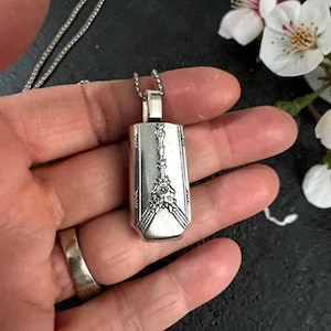 May include: Silver pendant necklace with a rectangular pendant. The pendant features a floral design and is suspended from a delicate chain. The pendant is approximately 1.5 inches long.