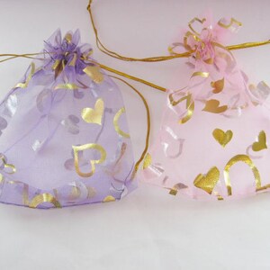 May include: Two sheer drawstring gift bags, one lavender and one pink, are displayed. Both bags are adorned with gold heart-shaped patterns. The bags are tied with gold ribbon and are suitable for small gifts or favors.