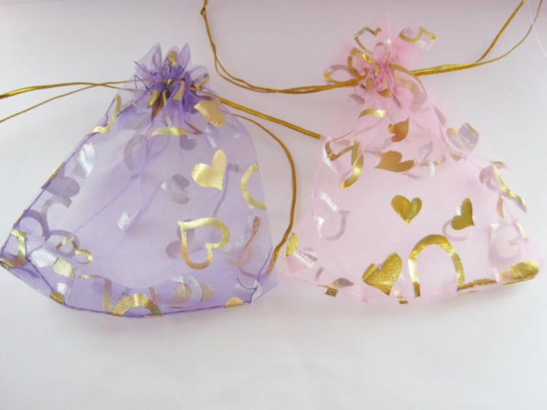 May include: Two sheer drawstring gift bags, one lavender and one pink, are displayed. Both bags are adorned with gold heart-shaped patterns. The bags are tied with gold ribbon and are suitable for small gifts or favors.