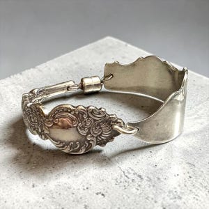 Puede incluir: Pulsera de plata hecha de cubiertos reciclados. Presenta detalles florales ornamentados y una cuchara decorativa. La pulsera está adornada con un cierre. Una joya única y original.