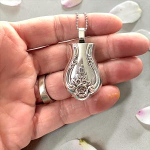 May include: Silver pendant necklace with a vase-shaped pendant. The pendant is decorated with embossed floral patterns. The necklace has a fine chain. The pendant is held in a hand, with a ring on the finger. Petal accents are in the background.