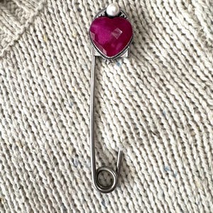 May include: A silver-toned safety pin brooch featuring a faceted, heart-shaped magenta gemstone at the top. The setting includes a small, round white pearl accent positioned directly above the heart, with intricate metal detailing around the stone.