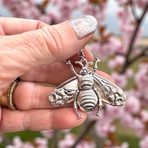 May include: A silver-colored bee pendant with detailed wings and body. The wings have a floral design. The bee is held in front of a blurred background of pink blossoms.