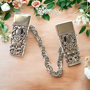 Puede incluir: Un par de broches de chal de metal plateado conectados por una cadena plateada. Los broches presentan un diseño floral ornamentado.