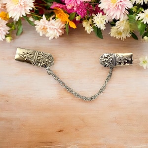 Könnte beinhalten: Ein Paar silberfarbene Schalschnallen, die durch eine silberne Kette verbunden sind. Die Schnallen haben ein florales Design.