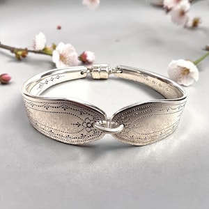 Puede incluir: Un brazalete de plata hecho con cucharas, con cierre magnético. El brazalete está hecho de cucharas reutilizadas, con patrones florales y punteados en los cuencos de las cucharas. Se muestra sobre una superficie gris con flores rosas en el fondo.