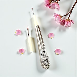 May include: Two seam rippers, one with a silver handle and floral design, the other with a white handle. Pink flower petals and a branch with pink flowers are scattered around the seam rippers.