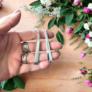 Peut inclure: Boucles d'oreilles argentées de forme rectangulaire allongée. Elles sont tenues dans une main, avec des fleurs en arrière-plan. Les boucles d'oreilles ont un dessus décoratif et une finition lisse et polie. Mesurent environ 10 cm de long.