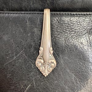 May include: A silver spoon with decorative patterns on the handle. The spoon is set against a dark, textured background, emphasising its intricate design and vintage style. The spoon's surface reflects light, showing its polished finish.