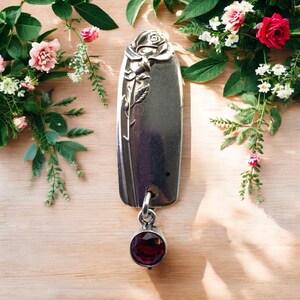 May include: A silver-toned key fob with a rose design at the top and a dangling, round, dark red gemstone. The key fob is rectangular with a floral design and a small hole for the gemstone. The background features flowers and greenery.